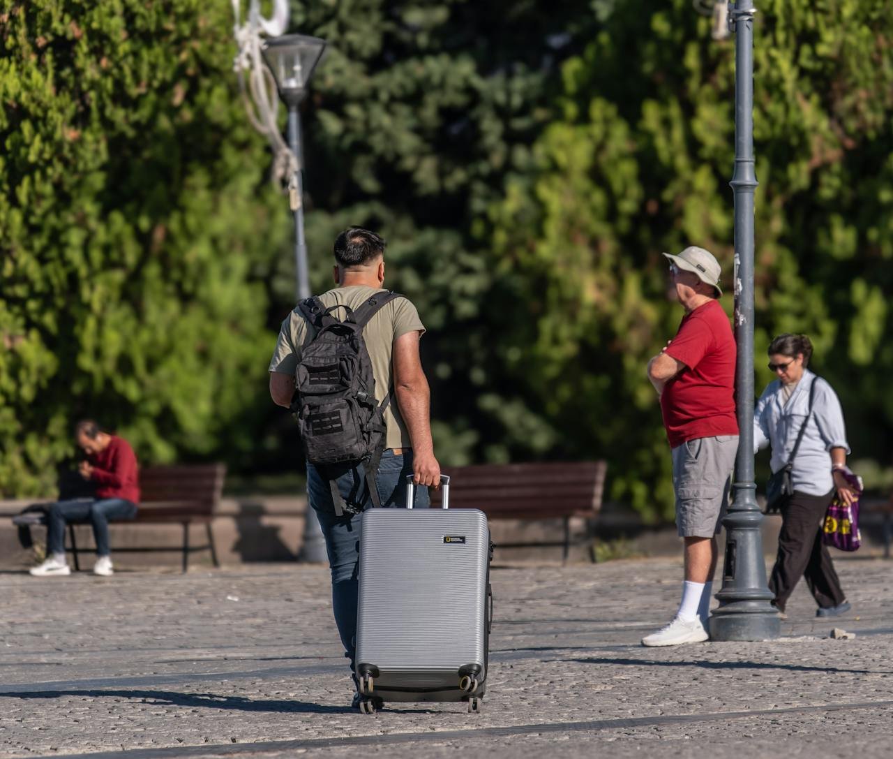 A man with a suitcase and backpack strolling in a sunny park in İzmir, Türkiye.